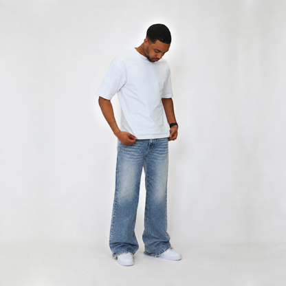 A man wearing a white crew-neck t-shirt, wide-leg light-wash blue jeans, and white sneakers, standing against a plain white studio background.