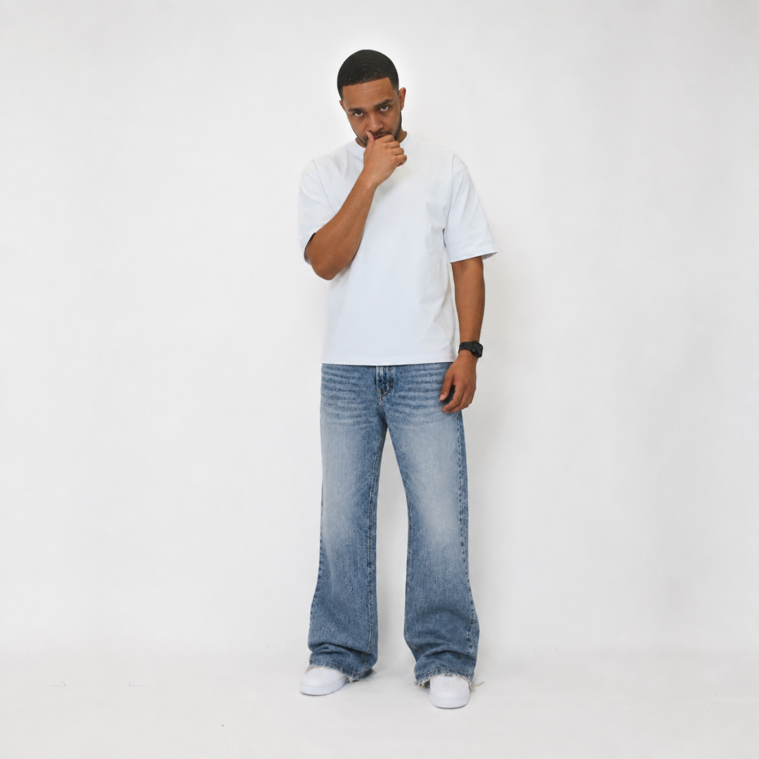 A man wearing a white crewneck t-shirt, wide-leg light blue denim jeans, and white sneakers, standing against a plain white studio background.