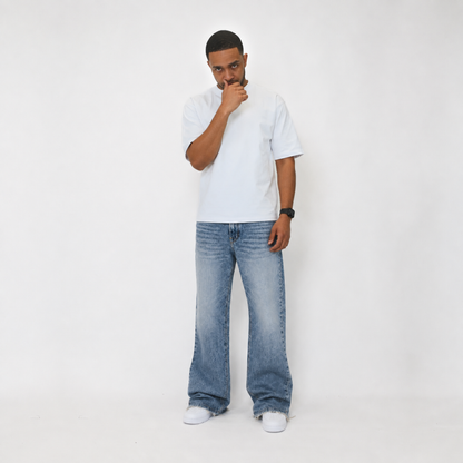 A man wearing a white crewneck t-shirt, wide-leg light blue denim jeans, and white sneakers, standing against a plain white studio background.
