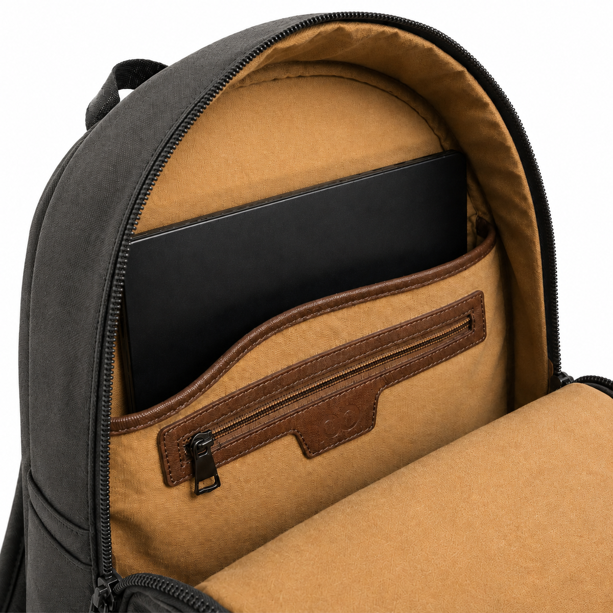 Close-up of the open interior of a dark gray backpack, showing a tan fabric lining, brown leather trim, a laptop compartment, and a zippered pocket holding a black laptop.